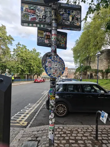 Photo of the back of a roadsign plastered in stickers, many of them featuring furry characters. Cars are parked in the background.