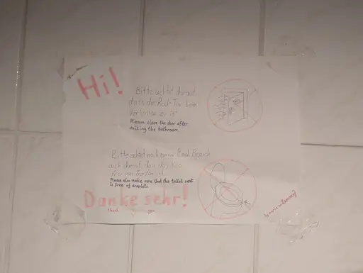 photo of a paper sheet taped onto a bathroom wall. it has text and drawings on it. the source text is German, but there are translations added. it reads "hi! please close the door after exiting the bathroom". a drawing of an open door with stinky lines coming out of it is crossed out, indicating it's unwanted. next "please also make sure that the toilet seat is free of droplets" and the drawing shows a toilet seat with droplets on it, being crossed out. there is an arrow pointing to the droplets. finally it says "thank you" with a heart below it. angled text on the tape says "by maria on lemmy"