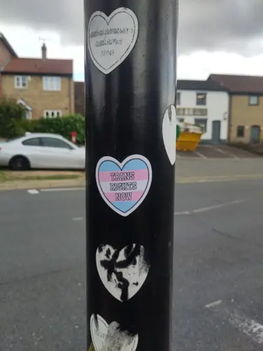 A photo of a traffic light pole with 4 worn/torn trans rights stickers and a fresh new one